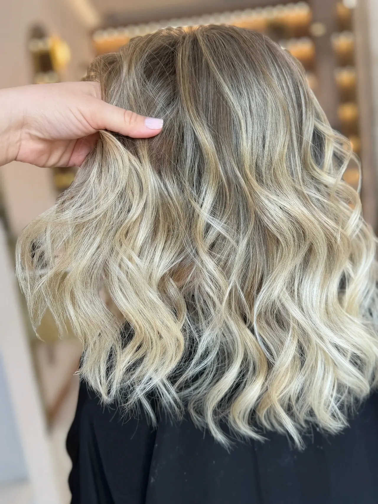 A stylist lifting softly waved blonde hair to reveal its texture and darker roots at Rové