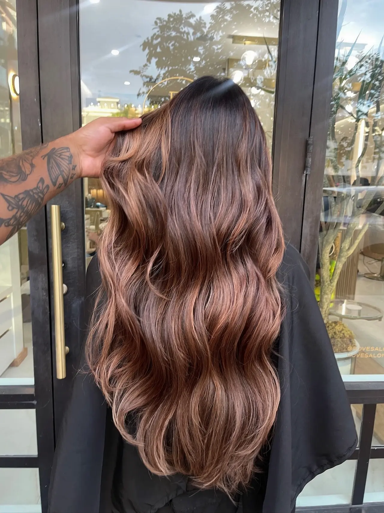 Long chestnut waves with warm highlights, shown from behind beside the salon’s glass doors