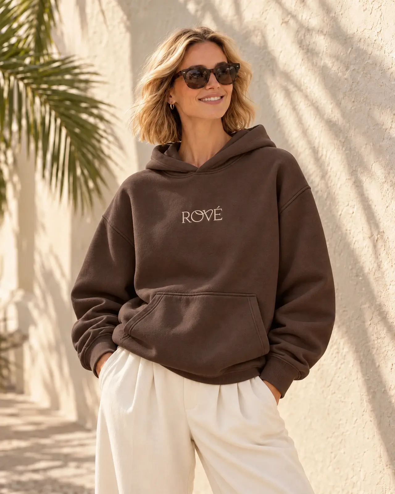 Blonde model wearing the espresso Rové hoodie with ivory trousers and sunglasses